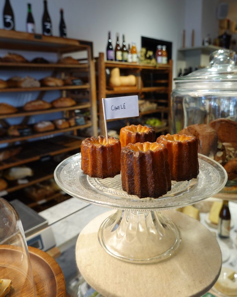 Vier canelé gebakjes zitten op een glazen taartstandaard met een klein "Canelé" vlaggetje in een bakkerij, met rekken brood en flessen op de achtergrond-een gezellig tafereel dat echte Genkse Gastvrijheid weergeeft, perfect voor een winterdate.