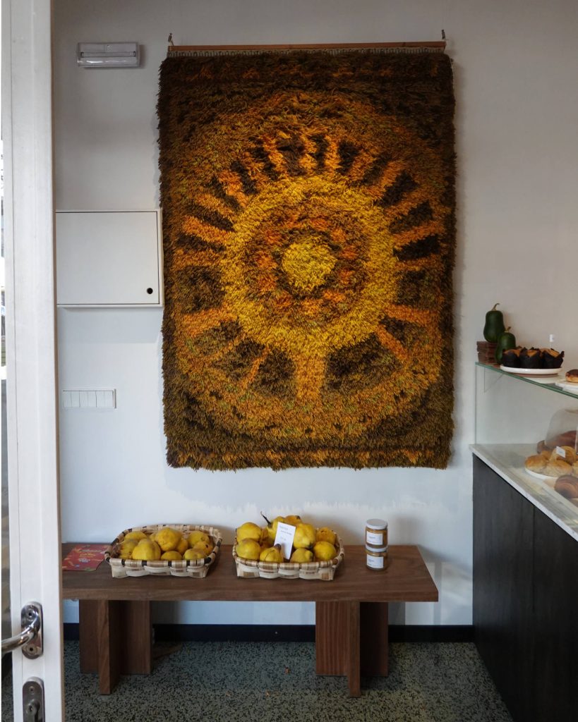 Een gezellig interieur straalt Genkse Gastvrijheid uit, met een gestructureerd, zonachtig wandkleed in warme gelen en bruintinten. Beneden staan twee manden met citroenen en een pot op een bankje. Rechts is een deel van een gebakvitrine te zien - een perfecte plek voor een winterdate.