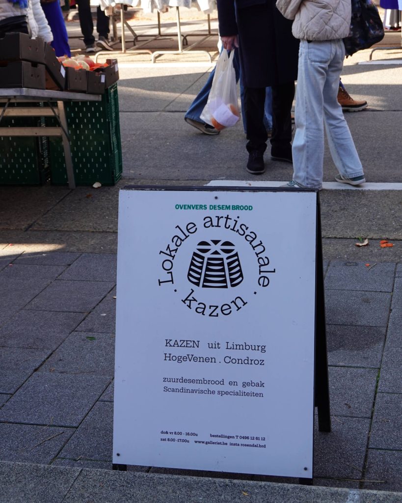Op een sandwichbord in een marktstraat staat "Lokale artisanale kazen" met een kaasgrafiek die de Genkse Gastvrijheid in de kijker zet. Het maakt reclame voor lokale kazen uit Limburg, HogeVenen en Condroz, plus zuurdesembrood en Scandinavische specialiteiten.
