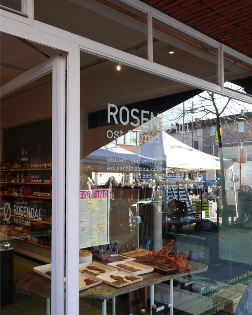 Op een winkelpui met grote ramen staat de naam "ROSENDAAL ost & brød". Binnen zijn rekken met goederen en houten planken met etenswaren te zien. Aan de overkant van de straat is een wintermarkt die Genkse Gastvrijheid uitstraalt met witte tenten en rondlopende mensen.