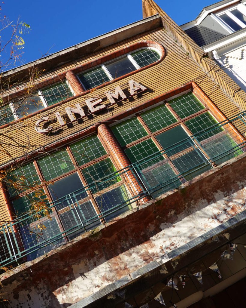 Een uitzicht vanuit een lage hoek op een oud gebouw met grote groene ramen en een verweerde gevel. "CINEMA" staat boven de ramen en nodigt bezoekers uit om te genieten van Genkse Gastvrijheid onder een strakblauwe hemel - perfect voor een gezellige winterdate.