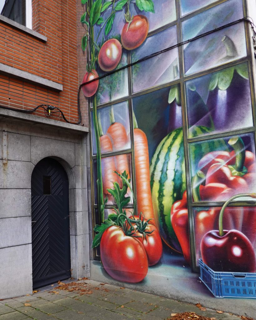 Een muurschildering op de zijkant van een gebouw in de geest van Genkse Gastvrijheid toont grote, kleurrijke groenten en fruit - waaronder tomaten, wortelen, watermeloen, aubergine en kersen - naast een donkere deuropening.