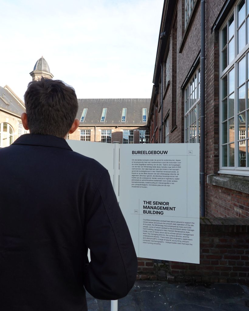 Een persoon met kort donker haar, van achteren gezien, leest een uithangbord over "The Senior Management Building" naast een bakstenen gebouw met grote ramen bij daglicht, een moment van Genkse Gastvrijheid vastleggend.