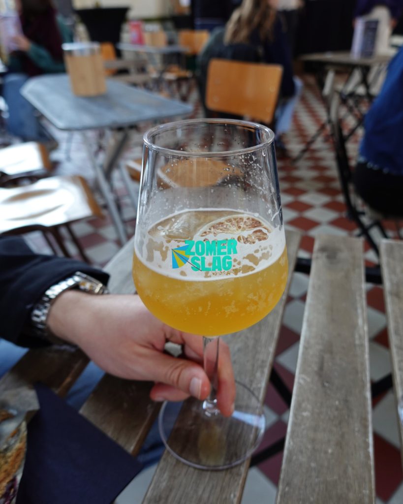 Een persoon geniet van een glas bier met het logo "Zomer Slag", echte Genkse Gastvrijheid aan een rustieke houten tafel binnen, omringd door geblokte tegelvloeren en houten stoelen - perfect voor een gezellige winterdate.