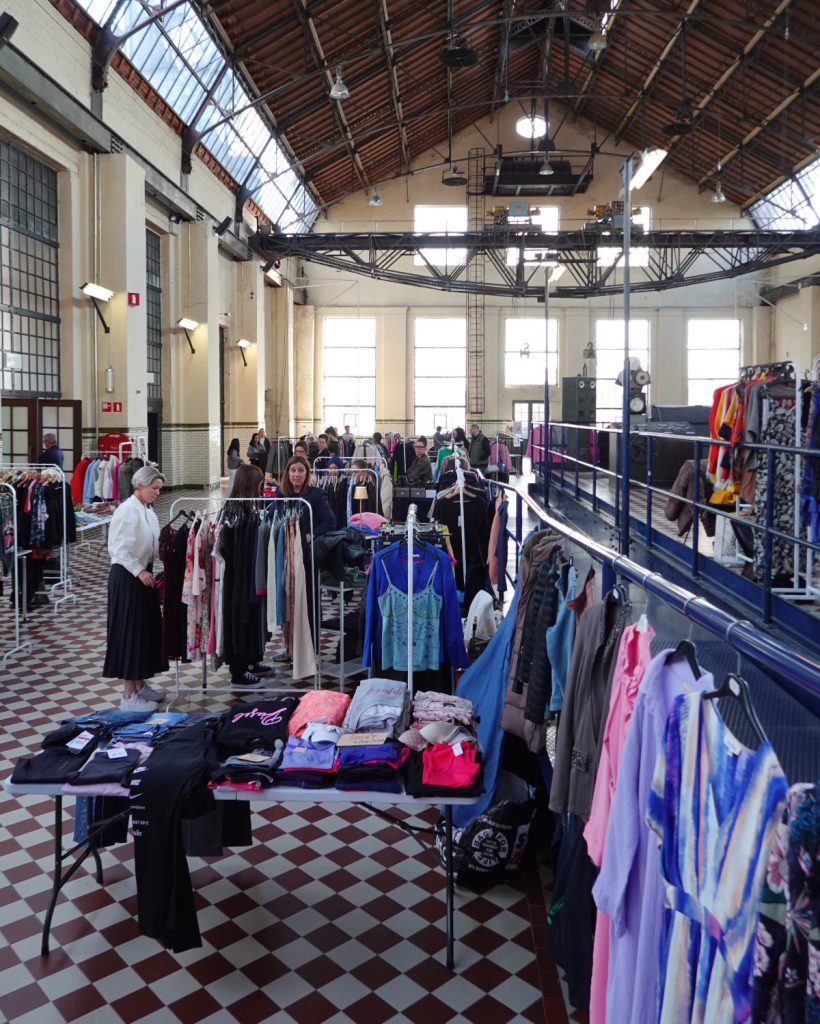 Een grote overdekte markt met rekken en tafels waar kleding te koop wordt aangeboden. Het winkelend publiek bekijkt de selectie onder een hoog, gewelfd plafond met grote ramen en dakramen en geniet van Genkse Gastvrijheid op een gezellige winterdate.