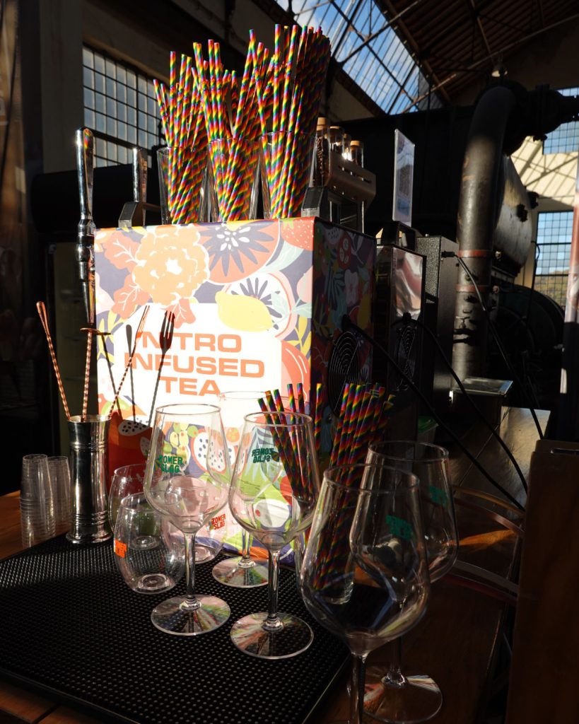 Een display met kleurrijke rietjes in een gebloemde "Nitro Infused Tea" doos, omringd door lege wijnglazen en bargereedschap, staat op een toonbank in een zonovergoten overdekte markt met industriële inrichting, die Genkse Gastvrijheid uitstraalt - perfect voor een gezellige winterdate.