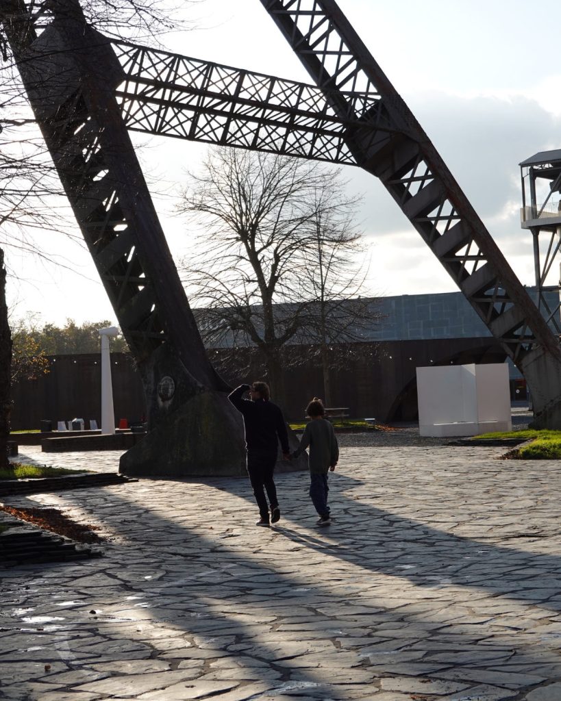 Twee mensen genieten van een winterdate, ze lopen hand in hand over een geplaveid pad onder grote metalen balken. Lange schaduwen strekken zich uit in het zonlicht terwijl bladerloze bomen en moderne gebouwen op de achtergrond een gevoel van Genkse Gastvrijheid oproepen.