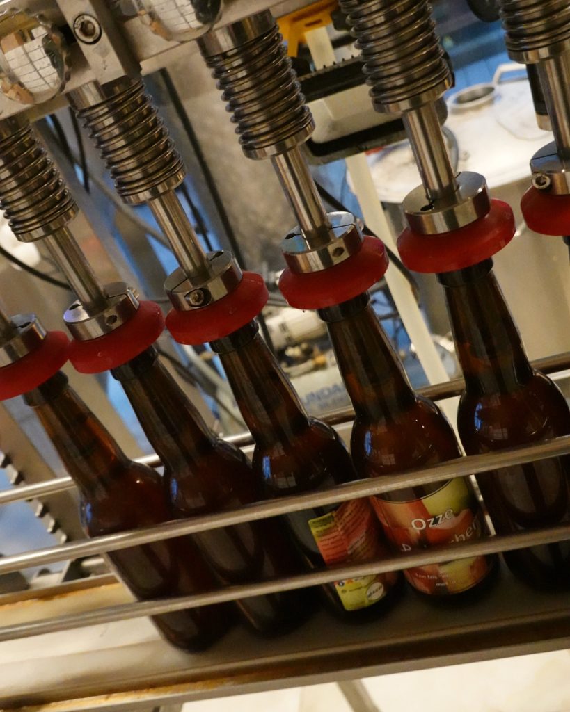 Close-up van een bottelmachine in Genk die bruine glazen bierflesjes vult met metalen sproeiers die vloeistof afgeven, met etiketten die Genkse Gastvrijheid en de warmte van een winterdag oproepen.