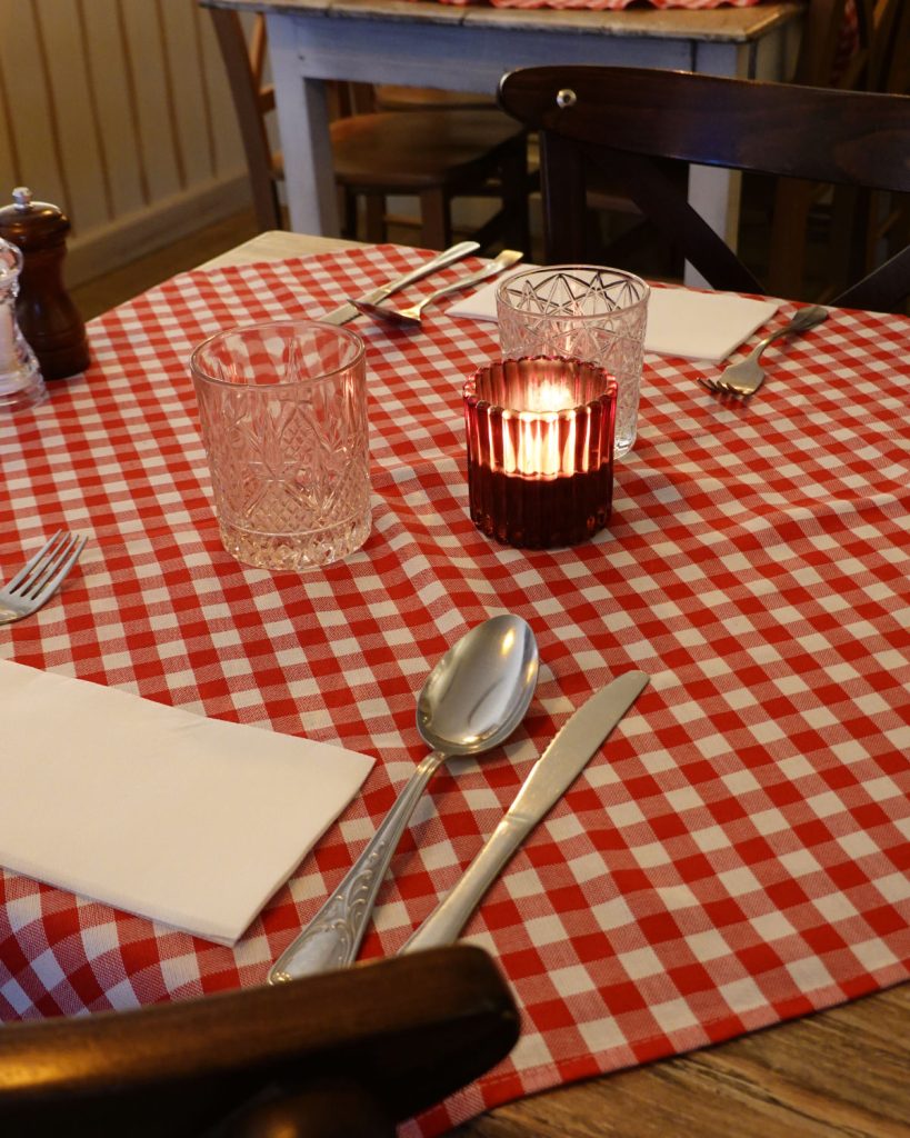 Een gedekte tafel met een rood en wit geruit tafelkleed, twee glazen, een kaars in een rode houder en bestek creëert een gezellige winterse sfeer, perfect om echte Genkse Gastvrijheid te ervaren.