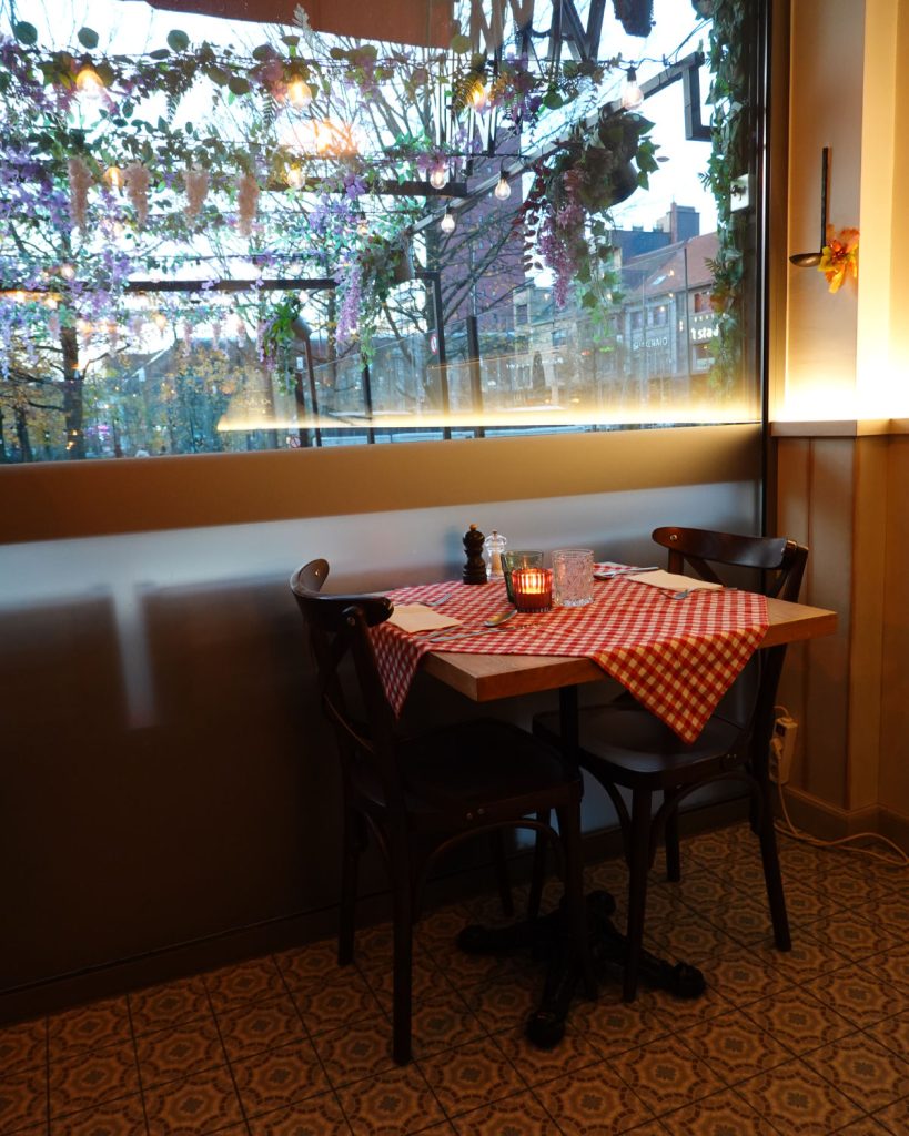 Een klein restauranttafeltje voor twee met een rood en wit geruit tafelkleed, een brandende kaars en specerijen staat naast een mat raam met planten en uitzicht op de straat buiten - perfect om te genieten van Genkse Gastvrijheid op een gezellige winterdag.