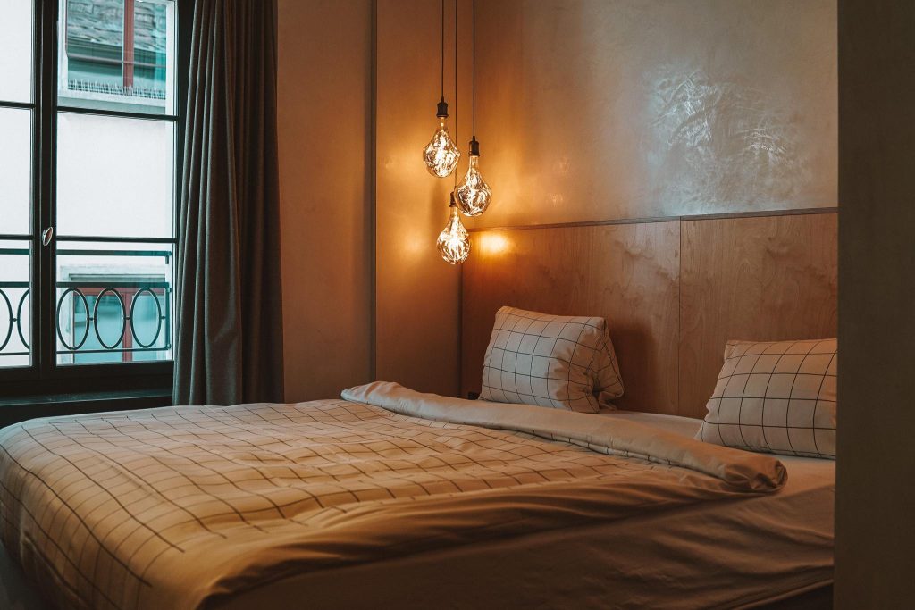 Gezellige slaapkamer met een tweepersoonsbed met een geruit dekbed en kussens, warme hanglampen boven het hoofdeinde en een raam met gordijnen die daglicht binnenlaten - perfect om te ontspannen na je herfst citytrip in Zwitserland.