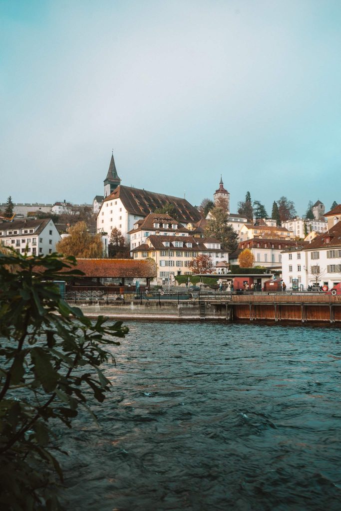 Een schilderachtig uitzicht op een Europese rivierstad met historische gebouwen, een kerk met een hoge torenspits en een klokkentoren, omlijst door groen onder een helderblauwe hemel - perfect voor een herfst stedentrip in Zwitserland.