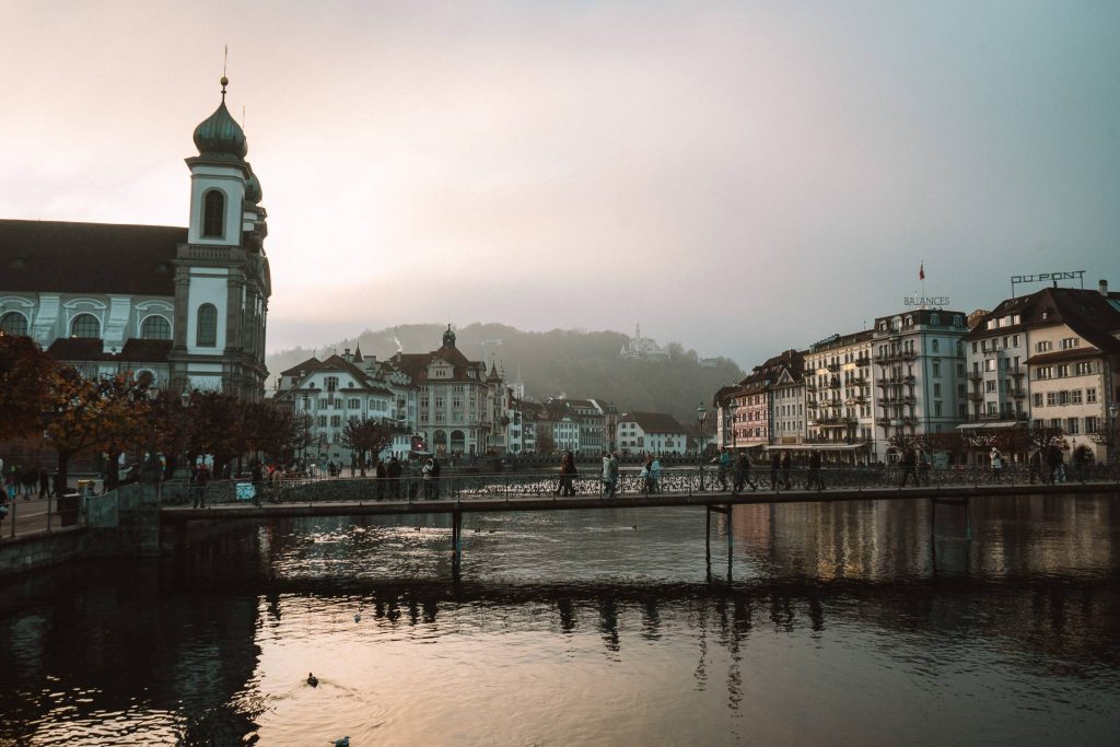 Een schilderachtig uitzicht op een Europese stad in Zwitserland met historische gebouwen, een kerk met een groene koepel, mensen die over een brug lopen en kalm water dat de stad weerspiegelt bij zonsondergang - een perfecte herfst citytrip bestemming met mistige heuvels op de achtergrond.