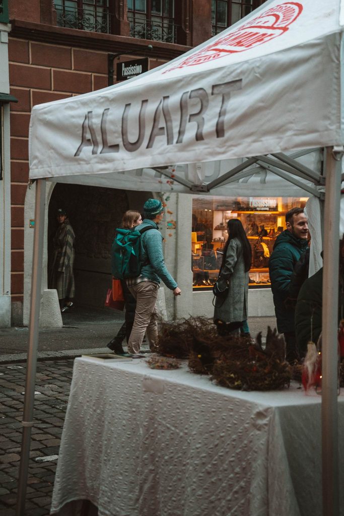 Een witte tent met "ALUART" erop staat op een openluchtmarkt tijdens een herfst citytrip in Zwitserland. Mensen lopen en kletsen langs de geplaveide straat, terwijl goederen worden uitgestald onder de tent bij een winkel met een verlichte etalage op de achtergrond.