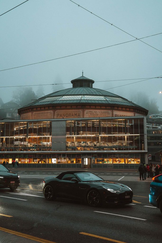Een rond, glazen gebouw met de naam "PANORAMA" straalt op een mistige avond tijdens een herfsttrip in Zwitserland, met auto's en mensen die langs de straat ervoor rijden.