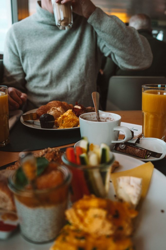 Een persoon in een grijze trui geniet van een ontbijt tijdens een herfsttrip in Zwitserland, nippend uit een glas aan een gedekte tafel met roerei, gebak, groenten, sap en een warme drank. De focus ligt op het heerlijke broodbeleg; de persoon is enigszins onscherp.