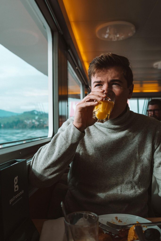 Een man met een grijze coltrui zit tijdens zijn herfsttrip in Zwitserland bij het raam van een trein of boot en drinkt sinaasappelsap. Voor hem op tafel ligt een menukaart met de tekst "gastronomie" en een gedeeltelijk opgegeten maaltijd.