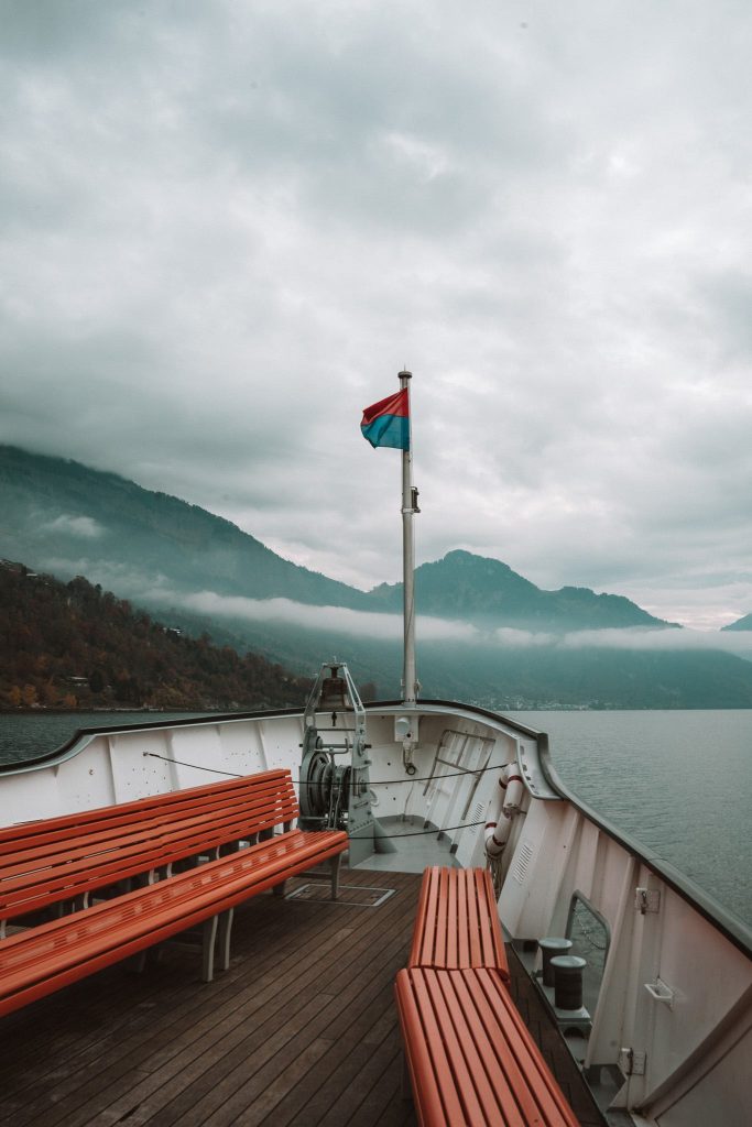 Een uitzicht vanaf het dek van een boot met lege houten banken, varend op een rustig meer omringd door bergen onder een bewolkte hemel; perfect voor een herfst citytrip in Zwitserland, met een roodblauwe vlag gehesen op de boeg.