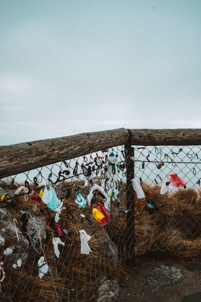 Een houten hek met gaas, versierd met kleurrijke linten, stroken stof en kleine voorwerpen, staat tegen een bewolkte hemel met uitzicht op een natuurlijk, graslandschap - een charmant voorbeeld van een herfstige citytrip in Zwitserland.