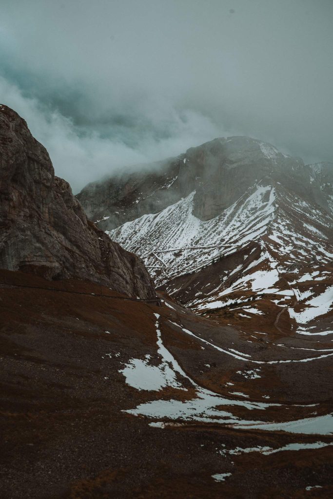 Besneeuwde bergen rijzen op door de mist en wolken, met rotsachtige hellingen en stukken sneeuw zichtbaar op de grond. De bewolkte lucht creëert een stemmige, dramatische alpine scène - perfect voor een herfst citytrip in Zwitserland.