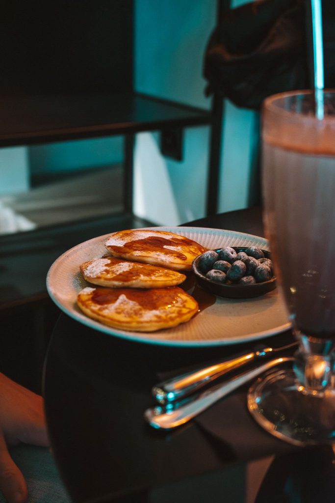 Een bord met kleine pannenkoeken met poedersuiker, bosbessen en een glas chocolademilkshake op een donkere tafel - de perfecte brandstof voor je herfsttrip in Zwitserland. Zilver bestek maakt het uitnodigende tafereel compleet.