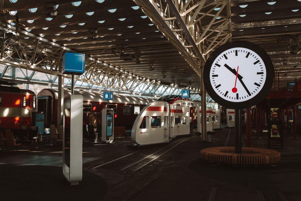 Een modern overdekt treinstation in Zwitserland, perfect voor een herfst citytrip, heeft verschillende witte treinen, digitale uithangborden en een grote ronde klok die 11:08 aangeeft terwijl het zonlicht door het dak met geometrisch patroon stroomt.
