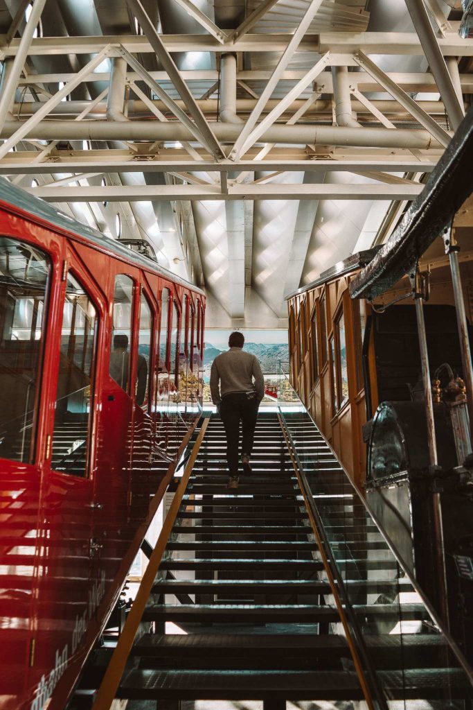 Een persoon loopt een glazen trap op tussen twee vintage treinwagons - een rode en een bruine - binnen een modern, industrieel gebouw met stalen balken en natuurlijk licht, dat de essentie van een herfst citytrip in Zwitserland vastlegt.