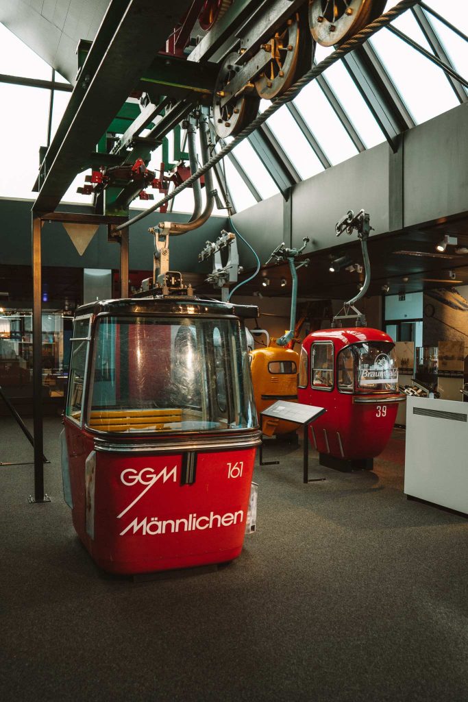 Een rode gondel met "Männlichen" erop staat binnen tentoongesteld, samen met andere kleurrijke gondelhutten, onder een railsysteem in een museumachtige omgeving - de perfecte inspiratie voor een herfst citytrip in Zwitserland.