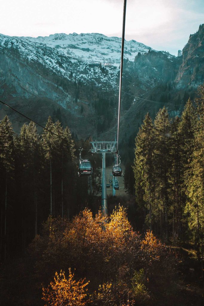 Kabelbanen glijden boven een bos met herfstkleurige bomen, perfect voor een herfst citytrip in Zwitserland. Besneeuwde toppen en ruige kliffen rijzen op in de achtergrond onder een gedeeltelijk bewolkte hemel.