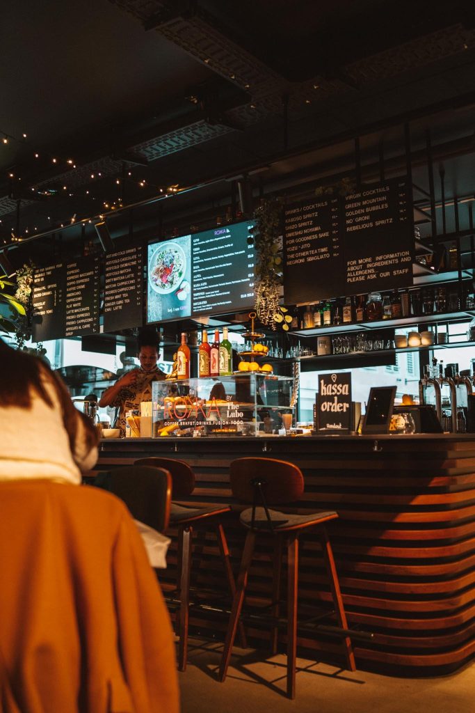 Een gezellige bar met warme verlichting, een barman achter de toonbank, drankjes en decor uitgestald, digitale menu's erboven en een persoon in een bruine jas zittend aan een tafel op de voorgrond.