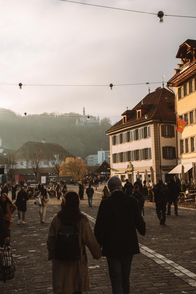Mensen lopen langs een geplaveide straat met traditionele Europese gebouwen bij zonsondergang, met bergen en een heuveltop zichtbaar op de achtergrond. Lichtsnoeren hangen boven het hoofd.
