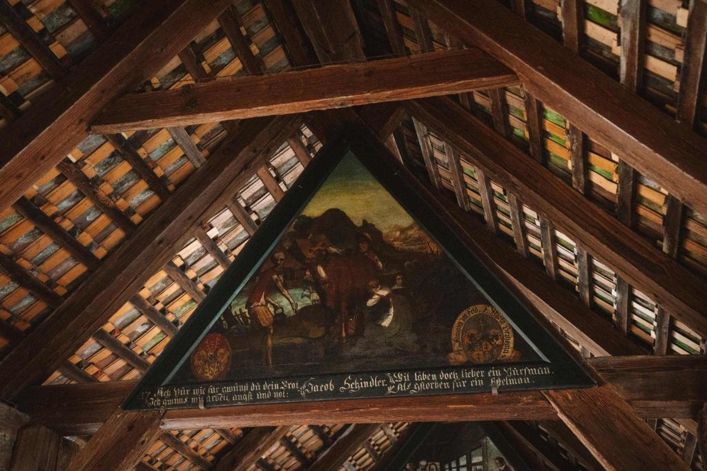 Een beschilderd driehoekig houten paneel met een historisch tafereel en Duitse tekst is gemonteerd onder de houten balken van een pannendak. Het licht valt naar binnen en accentueert de ingewikkelde details van het kunstwerk en het houtwerk.