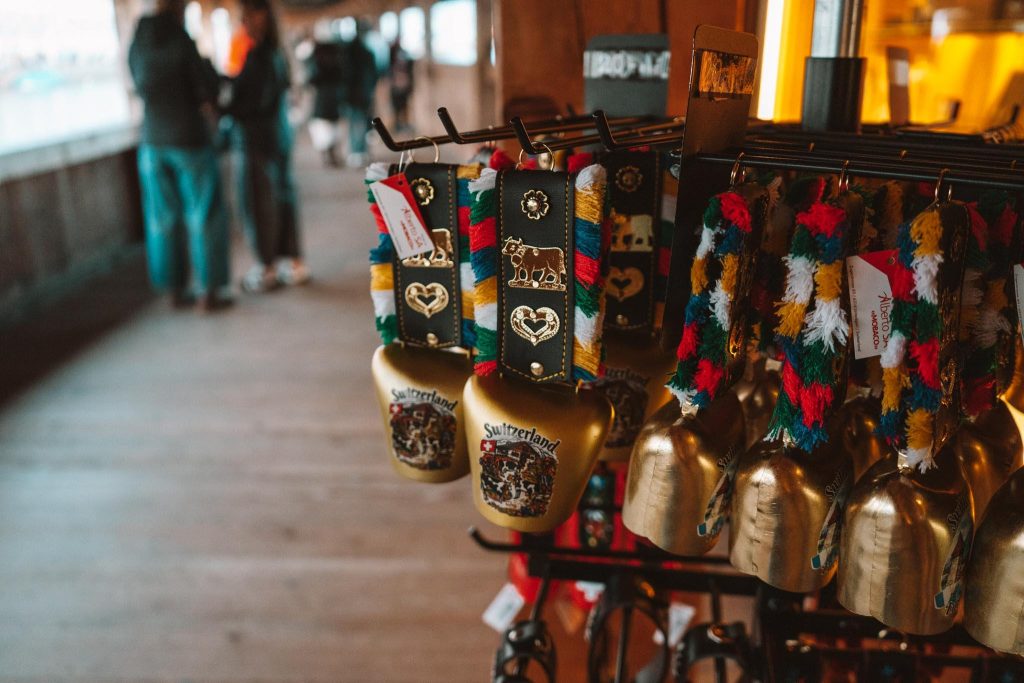 Verschillende decoratieve Zwitserse koebellen met kleurrijke riemen en Zwitserse emblemen hangen uitgestald in een houten overdekte loopbrug. Op de achtergrond staan twee mensen te praten bij de open kant van de loopbrug.