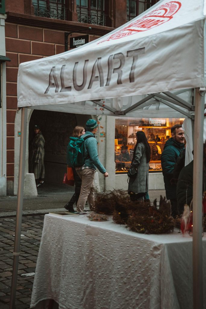 Een witte tent met het woord "ALUART" erop gedrukt staat op een straatmarkt. Mensen lopen voorbij en maken een praatje bij een etalage. De tafel onder de tent is bedekt met een doek en verschillende handgemaakte producten.
