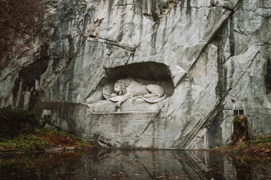 Een grote stenen sculptuur van een gewonde leeuw ligt uitgehouwen in een rotswand boven een stille, spiegelende vijver, omgeven door groen en rotsen. Het monument staat bekend als het Leeuwenmonument in Luzern, Zwitserland.