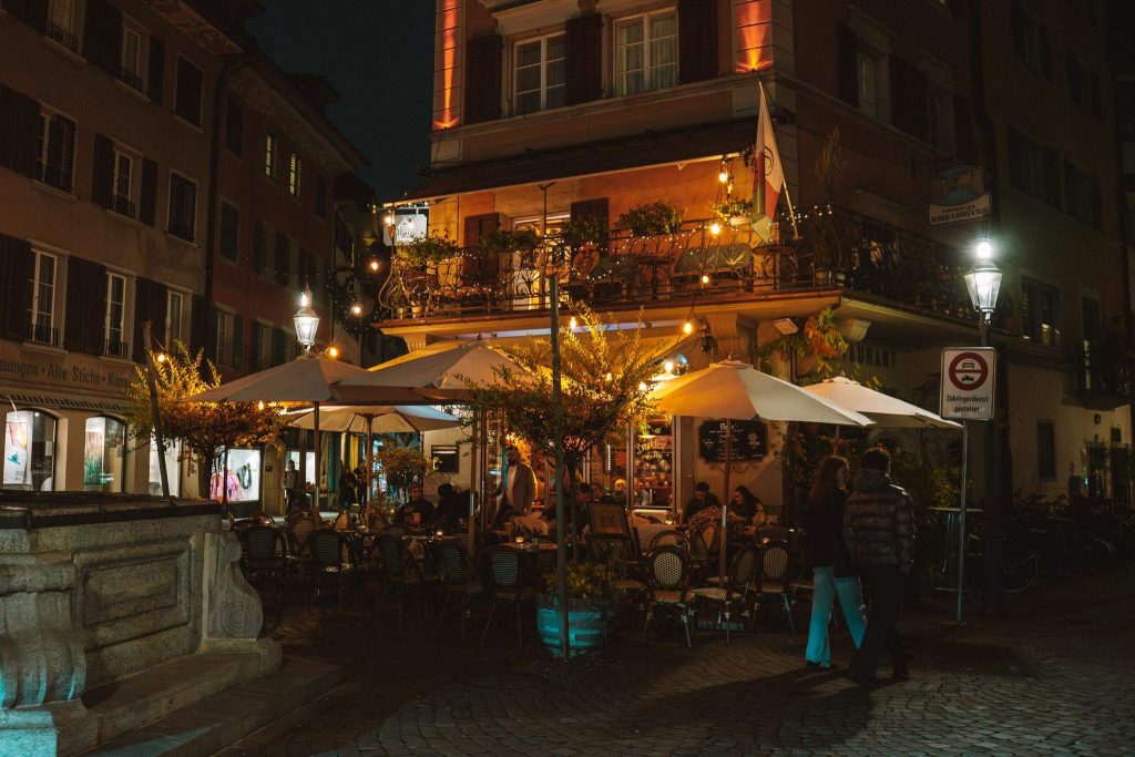 Een gezellig openluchtcafé 's avonds, met tafels en stoelen onder grote parasols, warme verlichting, etende mensen en een gebouw van twee verdiepingen met planten en balkons in een charmante Europese straatomgeving.