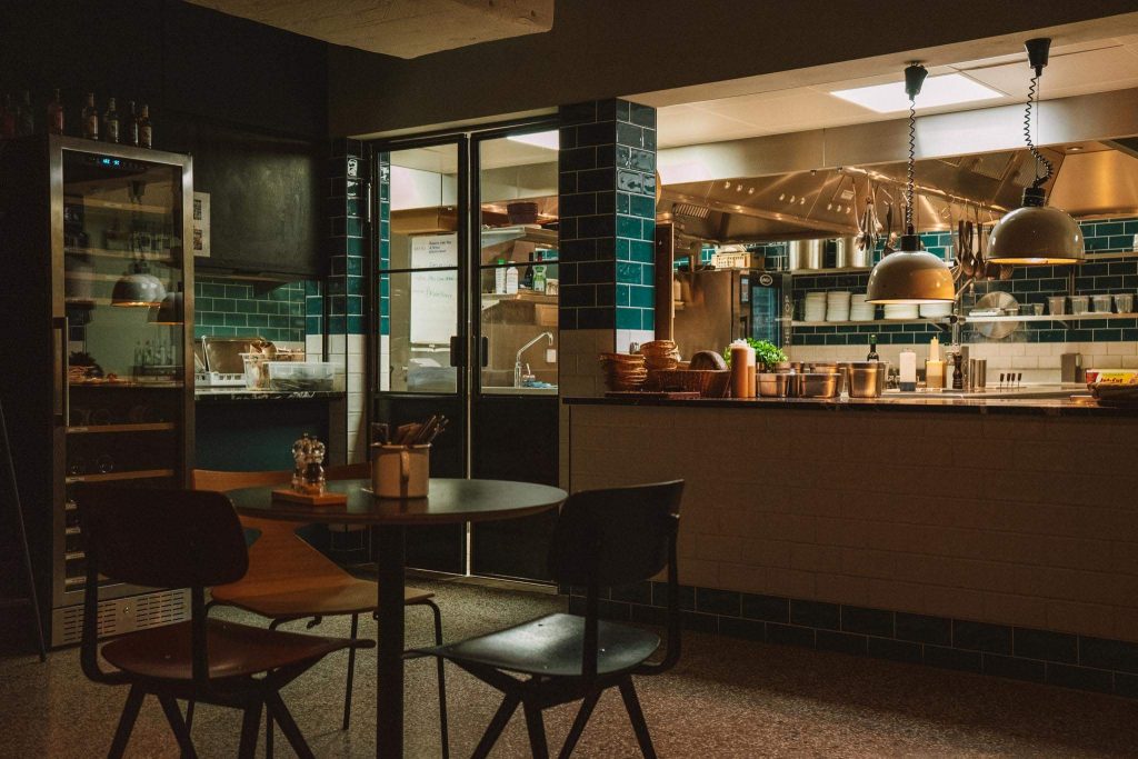 Een schemerig verlicht restaurantinterieur met een ronde tafel en twee stoelen op de voorgrond en een open keuken op de achtergrond met hangende lampen, planken met kookgerei en groenblauwe tegels op de muren.
