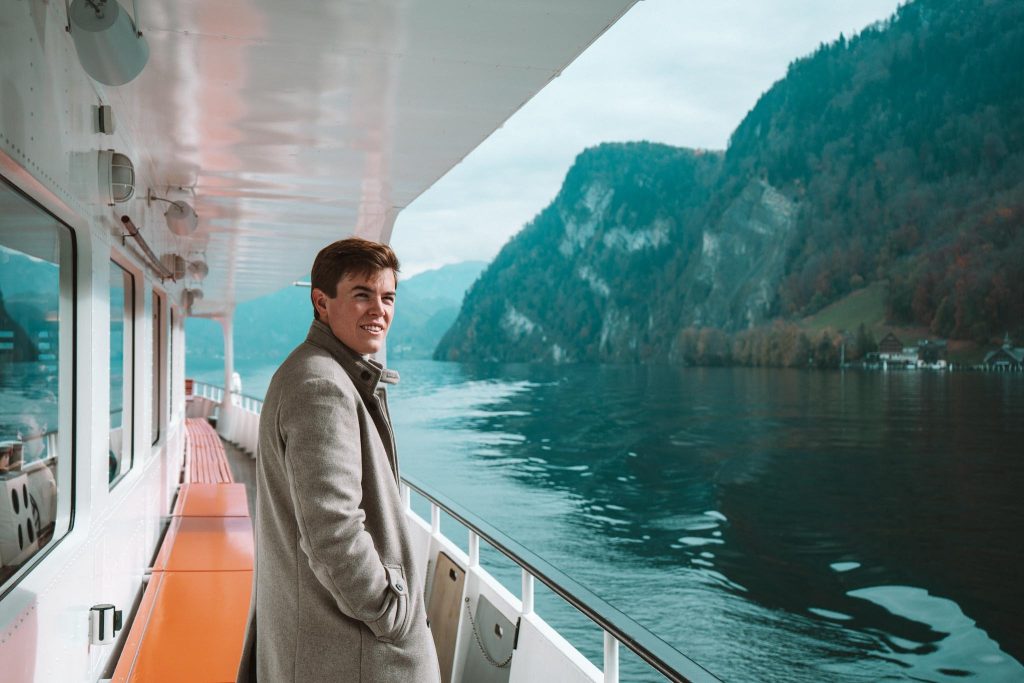 Een man in een lichtgrijze jas staat glimlachend op het dek van een boot. De boot ligt op een kalm meer omringd door groene, beboste bergen onder een bewolkte hemel.