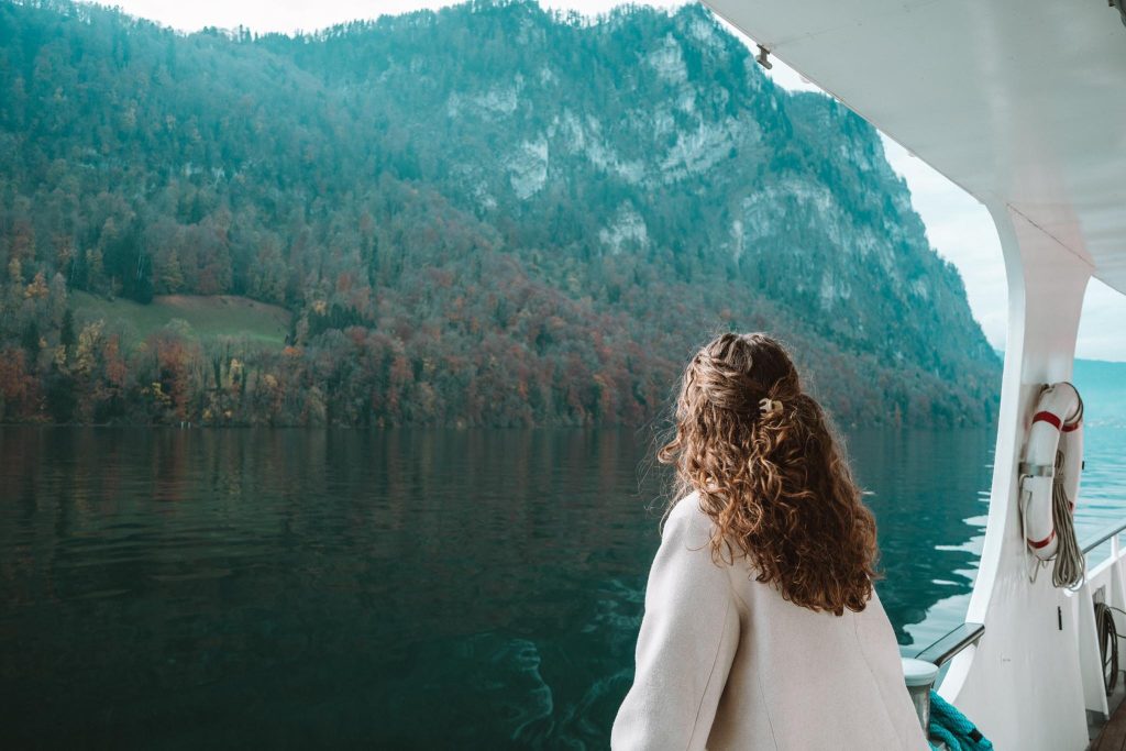 Een persoon met krullend haar, die een lichte jas draagt, kijkt vanuit een boot naar een kalm meer en een beboste berg bedekt met herfstbladeren. Het tafereel is vredig en sereen.
