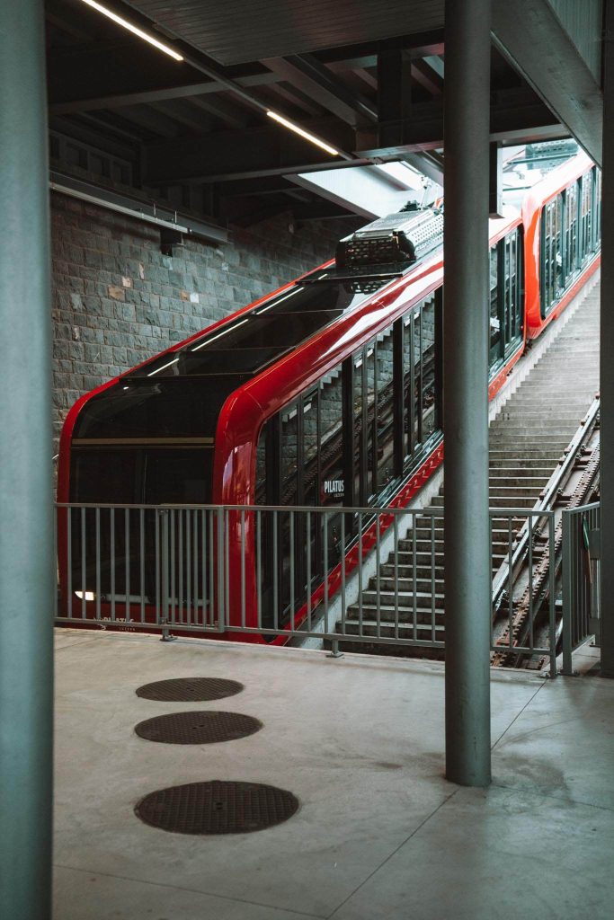 Een rode kabelspoorbaan staat geparkeerd op steile sporen in een modern station met betonnen vloeren, metalen leuningen en zichtbare structurele elementen. Het woord "Pilatus" staat op de trein.