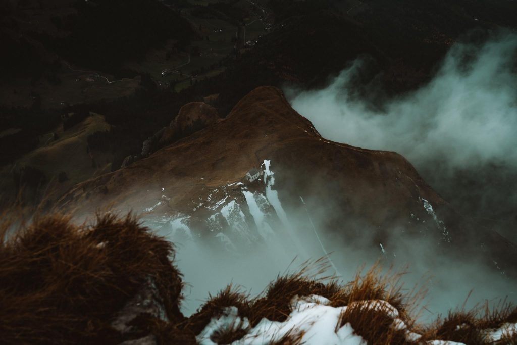 Een mistige bergrug met stukken sneeuw en bruin gras, gedeeltelijk verduisterd door wolken, van bovenaf gezien. De scène is dramatisch en sfeervol, met steile hellingen en een donkere, stemmige achtergrond.