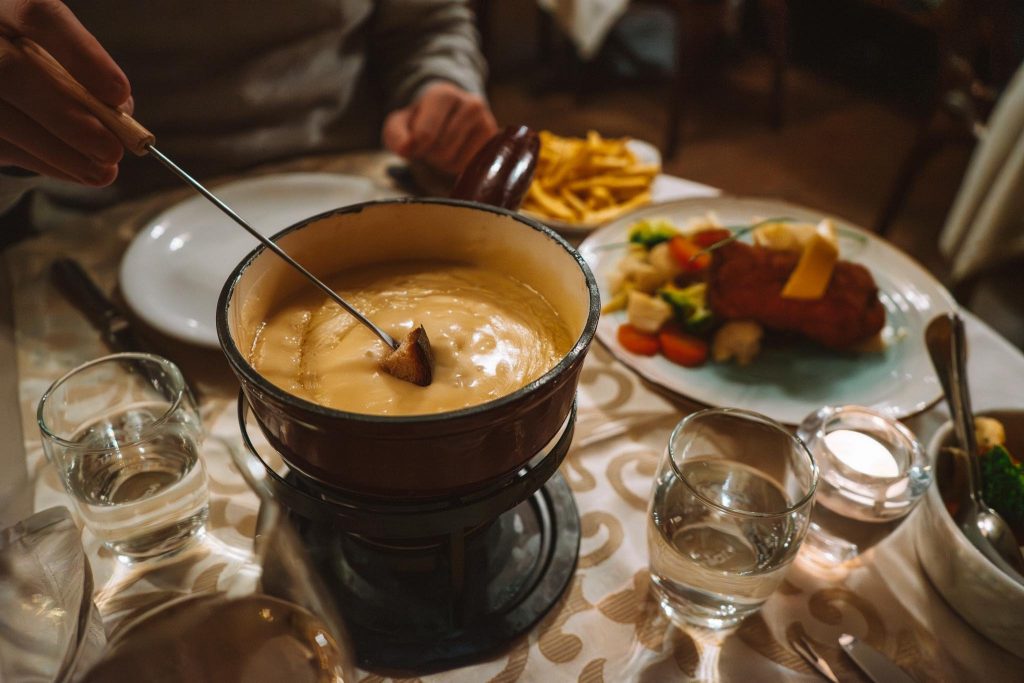 Een persoon doopt brood in een pot gesmolten kaasfondue op een restauranttafel met waterglazen, een bord friet en een bord vlees met groenten.