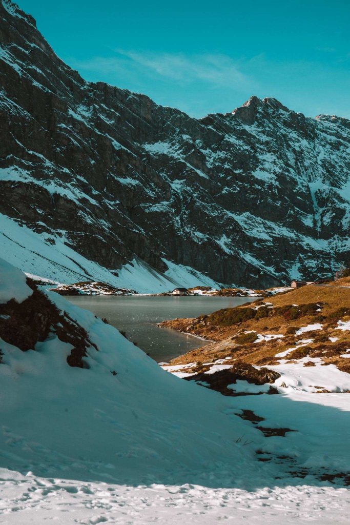 Een sereen berglandschap met besneeuwde hellingen, rotsachtige kliffen en een gedeeltelijk bevroren meer onder een helderblauwe hemel. Vlekjes groen gras zijn zichtbaar waar de sneeuw is gesmolten.
