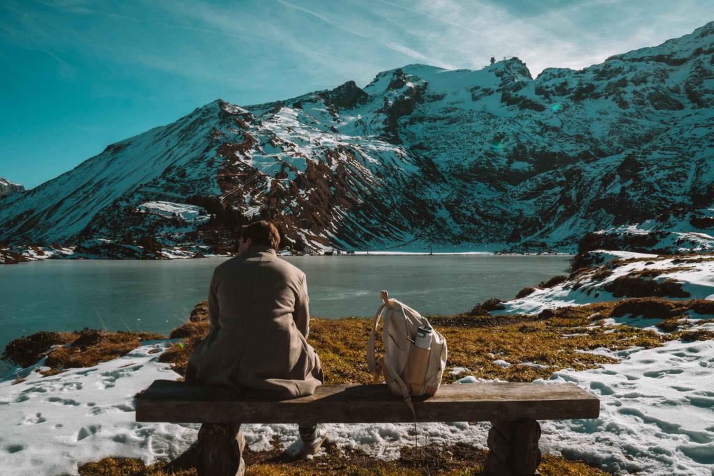Een persoon zit op een houten bankje tegenover een bevroren meer omringd door besneeuwde bergen, met een rugzak naast zich onder een strakblauwe hemel.