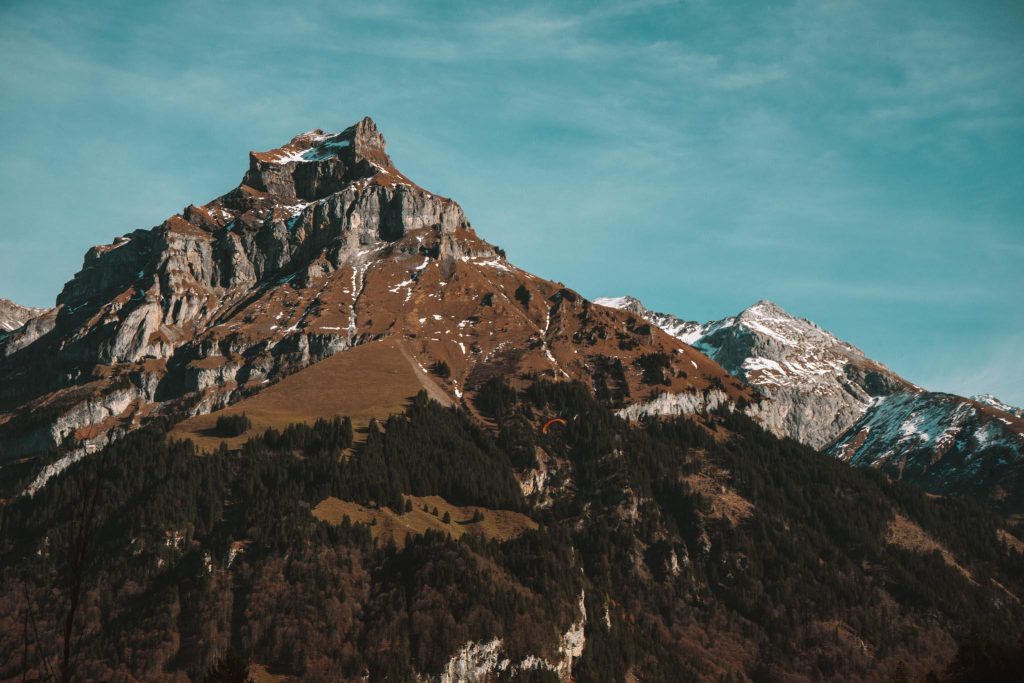 Grillige bergtoppen met stukken sneeuw steken boven beboste hellingen uit onder een blauwe hemel met pluizige wolken. Het ruige terrein toont een mix van rotsen, gras en bomen.