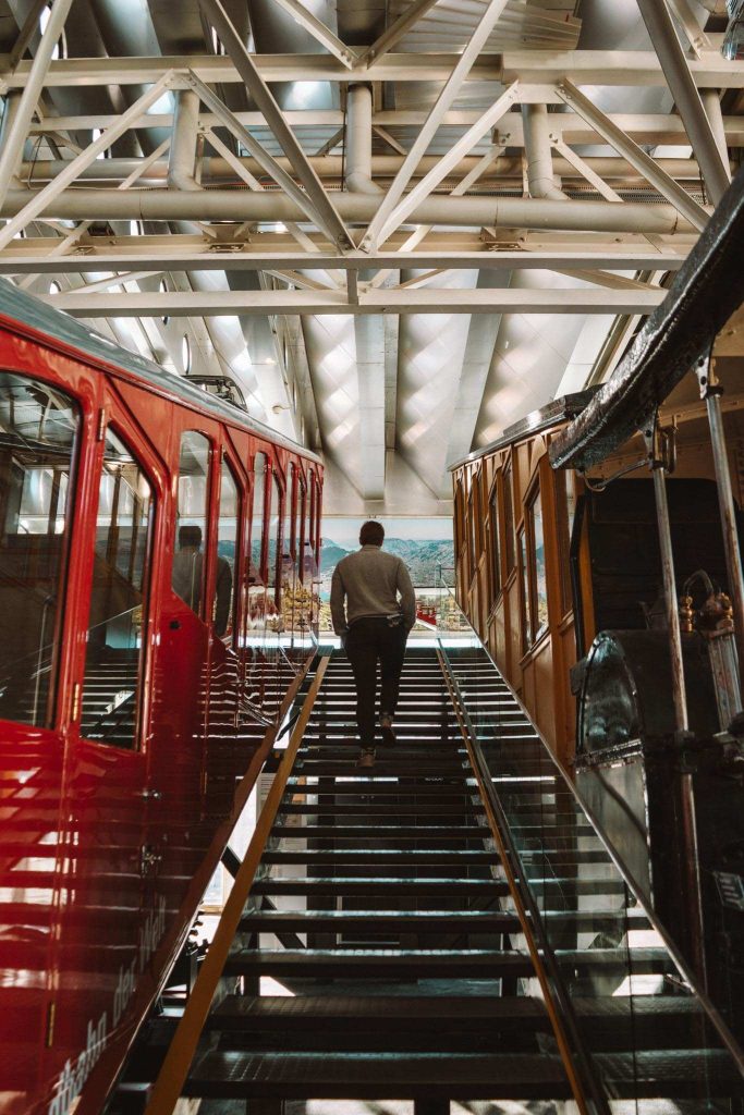 Een man loopt een glazen trap op tussen twee oude treinwagons - een rode en een gele - in een helder, industrieel gebouw met zichtbare balken en natuurlijk licht dat naar binnen stroomt.
