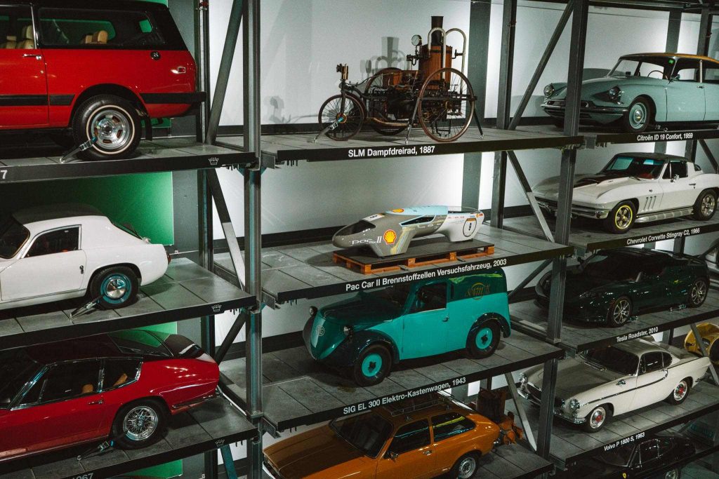 Een tentoonstelling van klassieke en vintage auto's op metalen planken in een museum, waaronder auto's in rood, groen, wit en groenblauw, evenals een oud stoomvoertuig en een futuristisch uitziende zilveren auto.