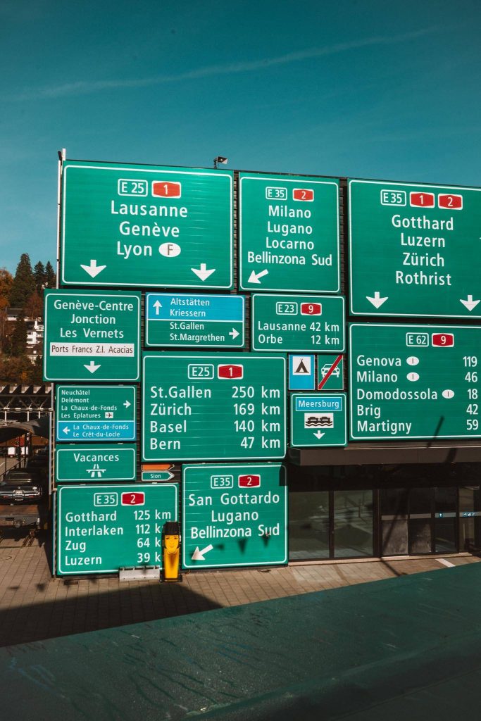 Een groep groene en blauwe Zwitserse verkeersborden met richtingen en afstanden naar steden als Lausanne, Genève, Milaan, Zürich, St. Gallen en Luzern, met een helderblauwe lucht op de achtergrond.