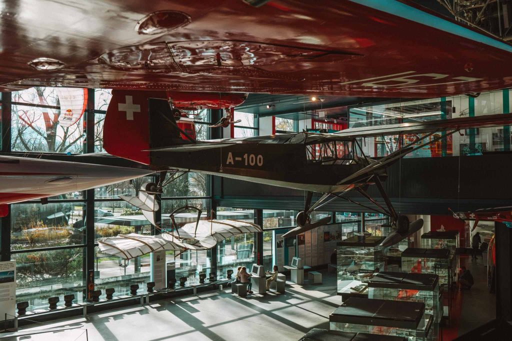 Een vintage vliegtuig met staartnummer A-100 hangt in een zonovergoten museum met grote ramen, met daaronder andere vliegtuigen en tentoonstellingen. Op de staart van het vliegtuig is een Zwitserse vlag te zien.