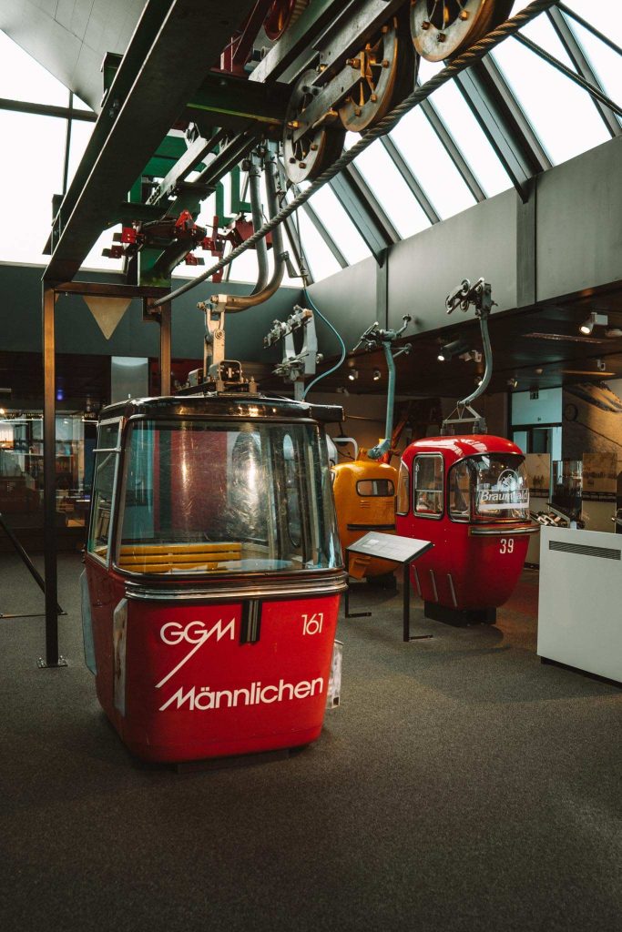 Oude gondelbanen die binnen worden tentoongesteld in een museum, met een rode baan op de voorgrond met het opschrift "GGM Männlichen 161" en andere op de achtergrond, onder een dakraam.